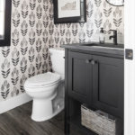 A black bathroom vanity cabinet, patterned wallpaper, toilet and wood flooring in a powder room.