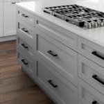 A gray shaker kitchen cabinet island with white quartz countertops and natural wood flooring.