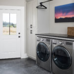 A laundry room with custom white storage cabinets, washer, dryer and a stained concrete floor.