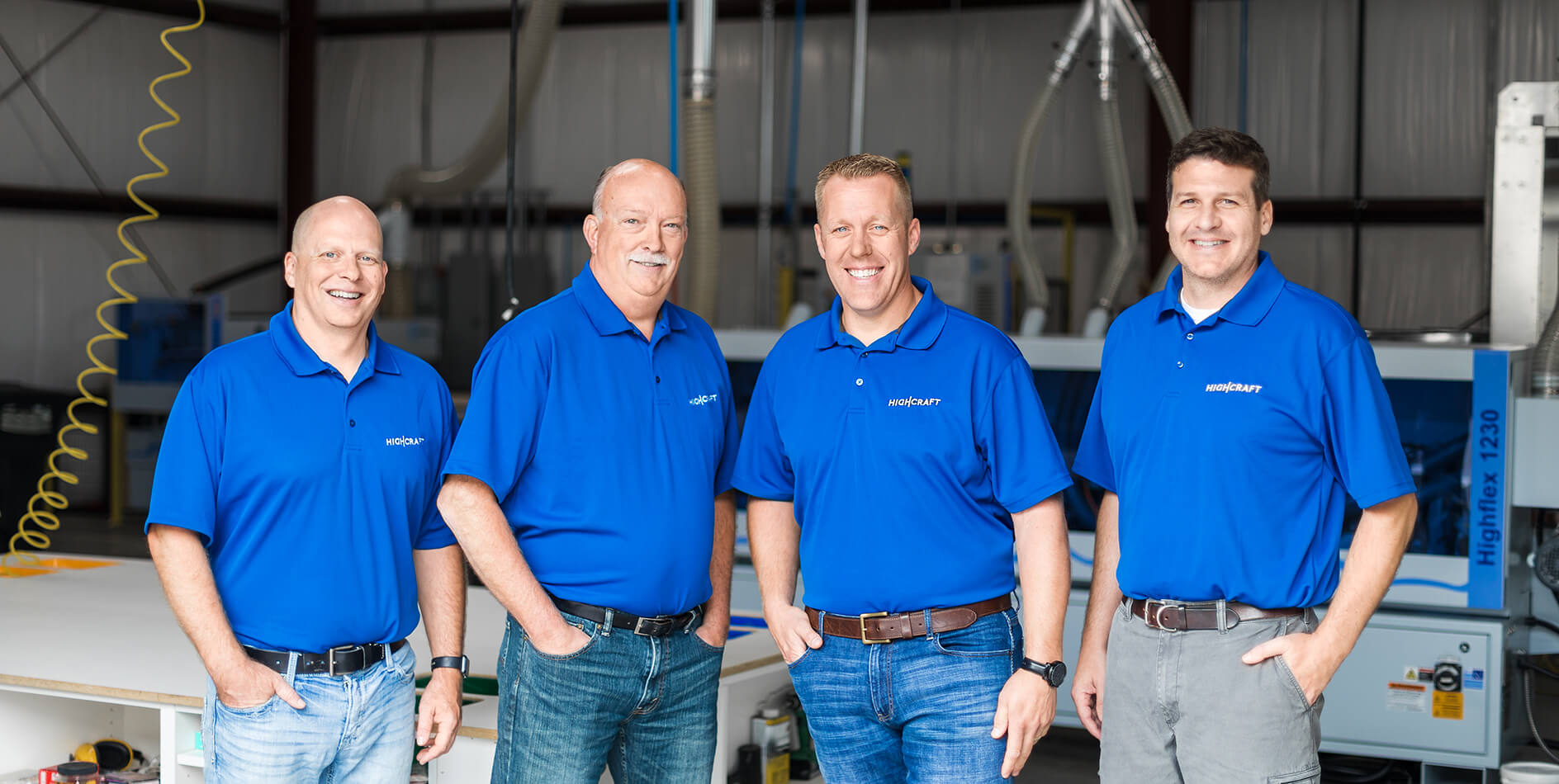 Four business partners in blue shirts standing by machinery in HighCraft’s cabinet manufacturing shop.